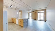 This thoughtfully designed urban loft offers a seamless open-plan layout that blends the kitchen and living areas into one expansive, light-filled space. The interior draws on an industrial aesthetic, showcasing exposed ductwork and piping along the ceiling, which adds character and a loft-like ambiance throughout. Warm wood-toned cabinetry lines the kitchen, complemented by a substantial center island with a striking granite countertop and built-in sink. Pendant lighting hangs above the island, providing focused illumination while enhancing the modern design. A dishwasher is integrated into the cabinetry for added convenience. The flooring transitions between wood-look planks in the kitchen zone and polished concrete-style flooring in the living area, reinforcing the industrial character of the space. Expansive windows and glass-paned double doors flood the interior with natural light and open onto a private balcony enclosed by a metal grid railing. From the balcony, elevated views stretch across a dynamic urban downtown landscape, offering a compelling backdrop of city rooftops and streets below. The overall atmosphere is contemporary, open, and urban — ideal for those who appreciate modern design with an industrial edge.