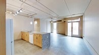 This thoughtfully designed urban loft offers a seamless open-plan layout that blends the kitchen and living areas into one expansive, light-filled space. The interior draws on an industrial aesthetic, showcasing exposed ductwork and piping along the ceiling, which adds character and a loft-like ambiance throughout. Warm wood-toned cabinetry lines the kitchen, complemented by a substantial center island with a striking granite countertop and built-in sink. Pendant lighting hangs above the island, providing focused illumination while enhancing the modern design. A dishwasher is integrated into the cabinetry for added convenience. The flooring transitions between wood-look planks in the kitchen zone and polished concrete-style flooring in the living area, reinforcing the industrial character of the space. Expansive windows and glass-paned double doors flood the interior with natural light and open onto a private balcony enclosed by a metal grid railing. From the balcony, elevated views stretch across a dynamic urban downtown landscape, offering a compelling backdrop of city rooftops and streets below. The overall atmosphere is contemporary, open, and urban — ideal for those who appreciate modern design with an industrial edge.