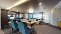 This contemporary amenity space is designed to foster social interaction and relaxation across a single, well-appointed floor. The layout is organized into several distinct functional zones, each with its own unique design character while maintaining a cohesive modern aesthetic throughout.

The lounge area features a generous sectional sofa paired with teal accent chairs, a sculptural chandelier overhead, and floor-to-ceiling glass walls that invite abundant natural light and views of the outdoor pool area. Sheer white curtains provide a soft contrast to the dark ceiling treatments and patterned carpet.

A stylish dining nook showcases a round table beneath a striking circular yellow ceiling medallion, creating a bold focal point. Adjacent to this is a secondary lounge with a teal-painted tray ceiling, warm cove lighting, brown leather chairs, and a mounted television, offering a more intimate gathering space.

The gourmet kitchen is a standout feature, equipped with stainless steel appliances including double wall ovens and a large refrigerator, dark wood cabinetry, a marble-topped island with teal bar seating, pendant lighting, and a three-dimensional tile backsplash.

The games area centers around a full-size billiards table, complemented by a herringbone wood accent wall, a wall-mounted display screen, and additional lounge seating. Large windows frame outdoor views, reinforcing the space's bright, connected atmosphere throughout.