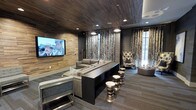 This stylish media lounge is designed for both comfort and visual impact. The room is immediately defined by its rich, dark wood-planked ceiling and complementary textured wood accent wall, which together create a warm, enveloping atmosphere. A large flat-screen television is mounted prominently on the wood feature wall, serving as the focal point of the space.

Seating is arranged in a tiered layout to optimize viewing angles, with plush gray upholstered sofas positioned along a raised platform. Metallic hourglass-shaped stools are scattered throughout, adding an elegant, contemporary touch. Near the window area, a wingback accent chair offers an alternative seating option alongside a round ottoman, creating a secondary lounge zone.

Floor-to-ceiling decorative curtains in a rich, marbled pattern frame the windows, adding texture and a sense of grandeur while filtering natural light. Recessed lighting in the ceiling provides ambient illumination throughout the space. The carpeted flooring in a neutral tone ties the design elements together cohesively.

Overall, the space balances modern design with warm, natural materials, creating an upscale, cinema-inspired environment ideal for entertainment and relaxation.