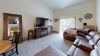 This single-floor residence offers a well-organized layout that balances comfort and functionality throughout its main living spaces. The living room is a standout area, featuring soaring vaulted ceilings that lend an open, airy feel to the space. A large sectional recliner sofa anchors the room, complemented by a media console with an electric fireplace insert. A sliding glass door floods the area with natural light and provides access to an outdoor balcony, while a staircase with wood and metal railing leads to a loft space above, adding architectural interest. The adjacent dining area features a round wood table beneath a chandelier, creating an inviting spot for meals. The galley-style kitchen is efficiently designed with warm oak cabinetry lining both sides, offering ample storage and counter space. White appliances, including a refrigerator and range, are paired with a double basin sink and tile flooring. The two bedrooms are carpeted and restful in character, each featuring bold wooden headboards with a rich, rustic finish and closet storage. The bathroom is finished with dark marble-style tile flooring, a dark wood vanity with a stone countertop, and a tub-shower combination, all accented with a cohesive nautical decorating theme throughout.