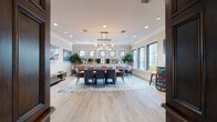 This single-floor space blends classic architectural character with modern interior design to create an inviting, multi-functional communal environment. The entrance foyer sets a refined tone with rich dark wood millwork, decorative columns, and arched brick accents framing a reception counter. Light hardwood-style flooring runs throughout, lending warmth and continuity across the space.

The central gathering area is anchored by a large wooden dining table surrounded by sleek gray upholstered chairs, positioned atop a layered area rug. Overhead, a striking industrial-modern chandelier with exposed Edison bulbs serves as a focal point. Crown molding and recessed lighting add a polished finish to the bright, airy interior.

Along the walls, additional seating arrangements featuring coral-accented chairs offer flexible social spaces, complemented by mirrored panels that visually expand the room. Tall fiddle-leaf fig trees introduce a natural element, while large windows along one side flood the space with natural light and views of the surrounding greenery.

A stylish lounge nook features a sculptural bar-height table with a vivid multi-colored base, white upholstered stools, and curated wall art. Adjacent to the entry, a corridor leads past a richly paneled elevator surround decorated with a colorful wall sculpture, reinforcing the space's thoughtful aesthetic throughout.