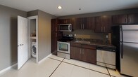 This single-floor residence is designed with a cohesive modern aesthetic, combining rich dark espresso cabinetry, granite countertops, and neutral gray walls throughout to create a polished and functional living environment. The kitchen serves as a standout feature, showcasing a full suite of stainless steel appliances — including an electric range, over-the-range microwave, dishwasher, and refrigerator — paired with dark granite countertops and a stainless steel sink. Crisp white tile flooring with contrasting inlay borders grounds the space and extends a clean, contemporary feel. Conveniently tucked into a dedicated alcove just off the kitchen is an in-unit washer, providing practical day-to-day functionality. The bathroom continues the design language established in the kitchen, featuring a dark wood vanity with matching granite countertop, an integrated sink, and a well-lit mirror above. White tile flooring with decorative border accents ties the rooms together seamlessly. The living area offers plush carpeting, soft neutral tones, and access to multiple rooms through clean white paneled doors. Natural light filters in through windows, brightening the space and adding an airy quality. The layout, as seen in the dollhouse view, is thoughtfully arranged for efficient flow between the kitchen, living, and bathroom areas.