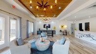 The Easterwood leasing office is a thoughtfully designed single-floor space that blends coastal charm with modern sophistication. The main lounge area commands attention with its elevated tray ceiling finished in warm natural wood planking, complemented by two striking brass-and-glass chandeliers that cast a warm, inviting glow throughout the room. Light-toned wood-look flooring runs seamlessly underfoot, enhancing the open, airy atmosphere.

The lounge is furnished with plush white slipcovered armchairs and a rounded stone-look coffee table, all anchored by a circular navy area rug. Shiplap-clad white walls add a fresh, transitional aesthetic, while large windows dressed in soft blue curtains allow abundant natural light and views of the surrounding greenery. French doors provide access to the exterior.

A built-in refreshment bar lines one wall, featuring sage-blue cabinetry with gold hardware, a beverage refrigerator, coffee station, and white quartz countertops beneath a wall-mounted flat-screen TV. A cozy dining nook with a round table sits nearby, ideal for casual meetings. The reception desk, boldly branded with the Easterwood name and a bird logo, serves as the central hub, with private office spaces accessible from behind it. The overall ambiance is clean, welcoming, and polished.