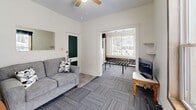 Located at 1002 Emerson Ave in South Bend, Indiana, this single-floor, one-bedroom, one-bathroom unit offers a practical and efficiently organized layout. The living room features gray carpet tile flooring, a ceiling fan, and a window with a roller shade that allows natural light to fill the room. The open sightline from the living room into the bedroom creates a connected feel throughout the space. The bedroom is a cozy, well-defined room with a large window overlooking the exterior, fitted with the same carpet tile flooring and a baseboard heater for climate control. The kitchen is straightforward and functional, equipped with a gas range, a utility sink, open shelving, and tile flooring — providing all the essentials in a no-frills layout. A cast-iron radiator adds a classic touch to the kitchen area. The bathroom rounds out the unit with wood-look vinyl flooring, a pedestal sink, a toilet, a mirrored medicine cabinet, and a tiled walk-in shower stall. Throughout the unit, white walls keep the interiors feeling bright and clean, while the consistent flooring choices and simple finishes contribute to a cohesive, utilitarian aesthetic well-suited for practical, day-to-day living.