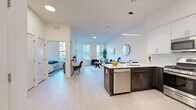 This single-floor residence is designed with a clean, contemporary aesthetic and an open-concept layout that seamlessly connects the kitchen, dining, and living areas. Light wood-toned flooring runs throughout, complementing the crisp white walls and creating a bright, airy atmosphere.

The kitchen is a standout feature, showcasing a two-tone design with dark lower cabinetry and white upper shaker-style cabinets. A quartz-topped island with bar seating provides both prep space and casual dining. Stainless steel appliances—including a gas range, built-in microwave, and full-size refrigerator—are complemented by a subtle tile backsplash and a linear pendant light above the island.

The living area is generously proportioned, featuring tall double-stacked windows that flood the space with natural light and create an open, expansive feel. The open floor plan allows for flexible furniture arrangements while maintaining a comfortable flow throughout.

The bedroom is well-sized, with multiple windows offering natural light and ample wall space. A closet with white panel doors provides practical storage.

The bathroom is finished with large-format stone-look wall tile surrounding a soaking tub and shower combination, paired with light floor tile, a white vanity, and a wide mirror, delivering a clean and spa-like finish.