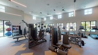 Located at 9260 Boca Vue Dr in Boca Raton, FL, this modern fitness center is thoughtfully designed to deliver a comprehensive workout experience in an open, airy environment. The space is defined by soaring ceilings with clerestory windows positioned high along the walls, flooding the interior with natural light and creating a spacious, energizing atmosphere. Large black-framed windows along the lower walls frame lush tropical views of palm trees and the surrounding grounds, enhancing the connection to the Florida outdoors.

The fitness center is well-appointed with a wide array of strength training equipment, including multi-station cable machines, weight benches, a full dumbbell rack, and resistance machines suited for a variety of muscle groups. Exercise balls and additional free weights are neatly organized in a designated functional training area. The cardio section features multiple treadmills with individual digital displays, lined up along windows overlooking the pool and landscape. Full-length mirrors amplify the sense of space throughout the facility.

The flooring is a striking two-tone rubberized surface in tan and gray, designed for durability and safety. Sleek linear pendant lighting and ceiling fans complement the contemporary aesthetic. Entry is through elegant glass doors with stainless steel hardware, accented by modern sculptural wall art, setting a refined tone from the moment of arrival.