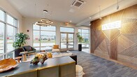 The Vibe at 3597 Paramount Dr in West Lafayette, Indiana, presents a thoughtfully designed lobby and hallway that together deliver a polished, contemporary first impression. The welcoming entrance is defined by large floor-to-ceiling windows and glass double doors that flood the space with natural light while offering open views of the surrounding landscape. A sculptural arrangement of circular ring pendant lights serves as a dramatic focal point overhead, complementing the neutral tones of the patterned carpet flooring below. The living area features a tufted leather sofa, eclectic seating, and a distinctive natural wood root coffee table, all anchored by a layered area rug. A vibrant moss wall art panel adds a refreshing organic element against the bright, white surroundings. The hallway transitions seamlessly from the lounge, showcasing a bold geometric wood-slat feature wall emblazoned with "The Vibe" branding, alongside a resident mailbox alcove. An adjacent wall clad in large-format white marble-effect panels frames a sleek black elevator door, adding a refined contrast. Potted greenery is thoughtfully placed throughout, enhancing the biophilic design aesthetic. The overall ambiance is modern, airy, and inviting.