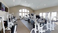 This fitness center is designed to accommodate a full range of workout needs within a bright, open environment. The space is defined by its striking vaulted ceiling, which rises dramatically overhead and is complemented by recessed lighting and a ceiling fan, ensuring the room remains well-lit and ventilated during use. Large arched windows along the front wall flood the interior with natural light, creating an inviting and energizing atmosphere.

The flooring transitions between carpet and rubber matting, with the matting strategically placed beneath the equipment to provide stability and protection. The fitness area is outfitted with an extensive selection of equipment, including elliptical trainers, treadmills, and a stair climber for cardiovascular workouts. A dedicated strength-training section features multiple weight machines and a dumbbell rack, catering to users at various fitness levels.

The neutral beige walls and clean layout contribute to a calm, focused environment. The generous ceiling height and wide-open floor plan allow for comfortable movement between equipment stations, making this fitness center both functional and aesthetically appealing.