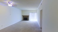 This single-floor residence offers a clean, functional layout designed for comfortable everyday living. The main living area is generously proportioned, featuring wall-to-wall carpeting that adds warmth underfoot and a sense of openness throughout the space. Crisp white walls and a smooth, light-colored ceiling enhance the bright, airy atmosphere, reflecting natural light effectively across the room.

A standout feature of the living area is the tile-surround fireplace, accented by a warm wood mantel, which serves as a natural focal point for the room. Positioned adjacent to the fireplace is a large sliding glass door fitted with vertical blinds, allowing for adjustable natural light and potential access to an outdoor area. The door's presence contributes significantly to the room's brightness and connection to the exterior.

The layout also includes a clearly defined entryway or passage along one wall, suggesting a logical and efficient flow between spaces. Crown molding details and ceiling vents point to a thoughtfully constructed interior with attention to finish quality. Overall, the space presents a neutral, move-in-ready environment with practical features, ample room for furnishing, and a welcoming ambiance anchored by the classic fireplace and natural light from the sliding door.