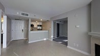 This single-floor residence offers a clean, contemporary design highlighted by a neutral gray and white color palette throughout. The open-concept layout seamlessly connects the living area to the kitchen, creating a spacious and functional flow. The kitchen is outfitted with white cabinetry, granite countertops, stainless steel appliances including a microwave and range, and a peninsula that doubles as a breakfast bar — ideal for casual dining or entertaining. Light wood-style flooring runs throughout the main living space, contributing to a bright and airy ambiance, while recessed lighting in the kitchen ensures the area is well-lit. A doorway from the living area leads into the bedroom, which features plush carpeting and a generous walk-in closet with built-in shelving, offering ample storage. The unit is equipped with central HVAC, visible through ceiling vents, ensuring year-round comfort. Clean white baseboards and smooth walls complete the modern, move-in-ready aesthetic. The overall design prioritizes practicality and understated elegance, making it a well-appointed and comfortable living space.