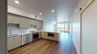 This thoughtfully designed single-floor residence offers a clean, contemporary aesthetic throughout. The open-concept kitchen is a standout feature, showcasing flat-front gray cabinetry extending to the ceiling, a white subway tile backsplash, light quartz countertops, and a full suite of stainless steel appliances including a refrigerator, range, dishwasher, and built-in microwave. Warm hardwood-style flooring runs seamlessly through the kitchen and living areas, complemented by recessed lighting overhead. A sliding barn door adds a modern architectural detail near the entryway corridor.

The living area opens toward a glass sliding door and large windows that invite abundant natural light and provide open outdoor views, leading to a private balcony. A dedicated bedroom is carpeted for comfort and is equipped with a ceiling fan; its oversized windows frame elevated views of the surrounding neighborhood and skyline, enhancing the airy feel of the space.

The full bathroom is finished with large-format tile flooring, a double-sink vanity with gray cabinetry and wide mirrors, and a combination soaking tub and shower with white tile surround. A hallway utility closet houses a stacked washer and dryer, providing in-unit laundry convenience. Crisp white walls and consistent finishes tie the entire residence together with a cohesive, modern sensibility.