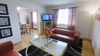 This single-floor space offers a comfortable and welcoming atmosphere, thoughtfully arranged to maximize both function and style. The living area serves as the heart of the home, anchored by a rich cognac leather sofa that adds warmth against the neutral-toned walls. A glass-top coffee table sits atop a bold black-and-white patterned area rug, providing a stylish focal point. A flat-screen TV is mounted on a media unit flanked by a bookshelf, offering both entertainment and storage. Natural light pours in through multiple windows dressed in elegant patterned curtains with a dark floral motif, creating a bright yet intimate ambiance. Baseboard heating units beneath the windows suggest year-round comfort. A decorative vase with autumnal floral arrangements adds a seasonal, homey touch. The living area opens seamlessly into a dedicated dining space, visible through an open entryway, where a wooden dining set rests on light hardwood flooring. The consistent hardwood flooring throughout ties the spaces together, contributing to a cohesive and warm aesthetic. Overall, the layout is practical and inviting, ideal for both relaxed everyday living and casual entertaining.
