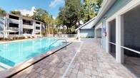 This outdoor pool amenity area offers a well-appointed retreat designed for relaxation and recreation. At its center is a generously sized swimming pool with crystal-clear blue water, flanked by a broad, paver-stone deck that provides ample space for lounging and socializing. Stainless steel entry rails make pool access convenient, and the surrounding fenced perimeter ensures a safe and defined environment.

Adjacent to the pool, a light blue-sided building houses a screened-in room accessible through full-length screen doors and large screened windows, allowing residents to enjoy the outdoors while staying sheltered from the elements. Inside this enclosed space, comfortable wicker seating and tropical greenery create a relaxed, lounge-like atmosphere with tile flooring throughout.

Thoughtfully curated landscaping enhances the grounds, with tall palms, mature trees, and fresh garden beds featuring tropical plantings integrated directly into the paver pathways. The canopy of surrounding trees provides natural shade, contributing to a serene and inviting ambiance. Umbrella-shaded seating areas are visible in the background, further extending the outdoor leisure offerings of this community pool space.
