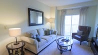 This single-story space showcases a well-appointed living area designed with comfort and style in mind. The room is finished in soft, neutral tones — pale green-grey walls and plush carpeting — creating a calm, welcoming atmosphere throughout. A large sliding glass door serves as a standout feature, allowing abundant natural light to fill the space while providing direct access to a private balcony, offering views of the surrounding exterior.

The living area is thoughtfully laid out, centered around a cushioned neutral sofa accented with decorative teal and green throw pillows. A round glass-top coffee table with a dark metal frame anchors the seating arrangement, adding a contemporary touch. A floor lamp provides warm ambient lighting, complementing the natural light from the balcony door dressed with sheer, floor-length curtains.

An ornately framed rectangular mirror mounted on the wall adds a classic decorative element and enhances the sense of depth and space within the room. Additional seating is partially visible, suggesting a generous and functional layout. Overall, the space balances modern furnishings with timeless accents, resulting in a bright, polished, and comfortable living environment.