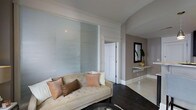 Located at The McCurdy, this single-floor residence showcases a thoughtfully curated interior design that balances modern sophistication with classic elegance. The living area is a standout feature, highlighted by floor-to-ceiling frosted glass sliding panels that serve as a striking architectural divider, diffusing light softly throughout the space while maintaining a sense of openness and privacy. Crown molding adds a refined finishing touch to the ceilings, complementing the warm taupe and white wall tones that define the overall color palette. Dark hardwood flooring grounds the living space, creating a rich contrast against the lighter walls and furnishings. A tufted cream sofa adorned with blush and copper-toned accent pillows anchors the seating area, conveying a sense of understated luxury. A floating mirrored console positioned against a feature wall adds depth and a contemporary accent, accompanied by a framed mirror and a touch of greenery that brings a subtle natural element indoors. The layout flows smoothly between spaces, with clean architectural lines and thoughtful design details that create a cohesive, polished ambiance throughout this distinguished residence.