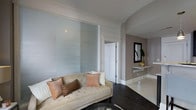 Located at The McCurdy, this single-floor residence showcases a thoughtfully curated interior design that balances modern sophistication with classic elegance. The living area is a standout feature, highlighted by floor-to-ceiling frosted glass sliding panels that serve as a striking architectural divider, diffusing light softly throughout the space while maintaining a sense of openness and privacy. Crown molding adds a refined finishing touch to the ceilings, complementing the warm taupe and white wall tones that define the overall color palette. Dark hardwood flooring grounds the living space, creating a rich contrast against the lighter walls and furnishings. A tufted cream sofa adorned with blush and copper-toned accent pillows anchors the seating area, conveying a sense of understated luxury. A floating mirrored console positioned against a feature wall adds depth and a contemporary accent, accompanied by a framed mirror and a touch of greenery that brings a subtle natural element indoors. The layout flows smoothly between spaces, with clean architectural lines and thoughtful design details that create a cohesive, polished ambiance throughout this distinguished residence.