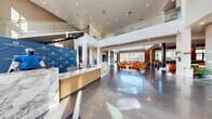 Located at 5050 W Russell Rd in Las Vegas, Nevada, the Otonomus Hotel lobby is a sophisticated, single-floor space spanning nearly 5,000 square feet of thoughtfully designed hospitality environment. The architectural design centers on soaring double-height ceilings and floor-to-ceiling glass windows that flood the interior with abundant natural light while offering open views of the surrounding outdoor landscape, including glimpses of palm trees and the Nevada sky.

The reception area greets guests with a striking marble front desk accented by brushed gold paneling, set against polished dark marble tile flooring that extends throughout the main lobby. A glass-railed mezzanine overlooks the space, adding vertical drama and dimension.

The lounge area is generously furnished with curved, modern seating in warm cognac, blush, and neutral tones, arranged around gold accent tables to encourage both social gathering and relaxed individual use. Curated artwork adorns the walls, contributing to the refined ambiance.

A dedicated dining alcove features arched wood-paneled booth seating with warm ambient lighting, while a stylishly appointed bar with stone cladding and hanging gold fixtures provides a sophisticated beverage service area with direct outdoor access. A bright convenience retail shop, "Essentials," rounds out the space with light wood flooring and well-stocked shelving, completing this polished, full-service hotel lobby experience.