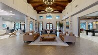 This expansive single-floor community space spans an impressively generous footprint, blending rustic architectural charm with modern functionality. The grand entrance sets the tone immediately, welcoming visitors into a soaring great room defined by dramatic vaulted ceilings with exposed wood beam trusses, elegant chandelier lighting, and natural stone columns that frame the open layout. Herringbone-patterned hardwood flooring flows throughout, anchoring comfortable leather seating arrangements around a substantial reclaimed wood coffee table. Clerestory windows invite abundant natural light, enhancing the airy, upscale ambiance.

Adjacent to the main living area, a spacious dining room features a large farm-style table and connects seamlessly to a casual kitchen and dining nook, where pendant lighting and bold teal accent walls add a contemporary pop of color. Compact bistro-style seating areas provide additional dining options in a relaxed setting.

A dedicated living room with custom teal built-in cabinetry and shelving serves as a comfortable media lounge. The well-appointed office area offers individual workstations separated by paneled dividers, providing privacy and focus, alongside a private conference room adorned with bold artwork.

The fitness center is fully equipped with strength and cardio machines, mirrored walls, rubber flooring, and ceiling fans, delivering a complete exercise environment. Together, these thoughtfully designed spaces create a cohesive, welcoming, and highly functional facility.