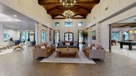 This expansive single-floor community space spans an impressively generous footprint, blending rustic architectural charm with modern functionality. The grand entrance sets the tone immediately, welcoming visitors into a soaring great room defined by dramatic vaulted ceilings with exposed wood beam trusses, elegant chandelier lighting, and natural stone columns that frame the open layout. Herringbone-patterned hardwood flooring flows throughout, anchoring comfortable leather seating arrangements around a substantial reclaimed wood coffee table. Clerestory windows invite abundant natural light, enhancing the airy, upscale ambiance.

Adjacent to the main living area, a spacious dining room features a large farm-style table and connects seamlessly to a casual kitchen and dining nook, where pendant lighting and bold teal accent walls add a contemporary pop of color. Compact bistro-style seating areas provide additional dining options in a relaxed setting.

A dedicated living room with custom teal built-in cabinetry and shelving serves as a comfortable media lounge. The well-appointed office area offers individual workstations separated by paneled dividers, providing privacy and focus, alongside a private conference room adorned with bold artwork.

The fitness center is fully equipped with strength and cardio machines, mirrored walls, rubber flooring, and ceiling fans, delivering a complete exercise environment. Together, these thoughtfully designed spaces create a cohesive, welcoming, and highly functional facility.