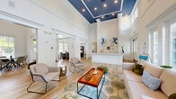 Located in Tampa, FL, this single-floor commercial space offers a thoughtfully designed layout that balances openness with functional, defined work areas. The centerpiece of the space is a dramatic great room featuring soaring double-height ceilings accented by a striking deep-blue coffered ceiling detail with recessed lighting — a bold architectural element that adds character and visual impact. Light wood-look flooring runs throughout, complementing the crisp white walls and trim for a clean, contemporary aesthetic.

Floor-to-ceiling windows and French doors line the main lounge area, flooding the space with natural light and offering views of the lush Florida landscape outside. The lounge is furnished as an inviting gathering area with comfortable seating, a coffee table, and accent pieces that contribute to a welcoming ambiance.

A reception counter anchors the central zone, with an open hallway connecting to private offices. These windowed offices are well-lit and functional, each accommodating a full workstation setup. A dedicated meeting room with a round dark-wood table and seating provides a professional setting for discussions, with natural light filtering through slatted blinds. A service counter with a coffee station adds convenience. A bathroom and entrance area complete the layout, making this space well-suited for professional or service-oriented use.