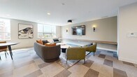 Located at 1110 Lawson Avenue in Kelowna, British Columbia, this well-appointed amenity lounge offers a thoughtfully designed single-floor space that blends comfort and modern aesthetics seamlessly. The open-concept layout accommodates both a living area and a kitchenette, creating a versatile environment suited for relaxation and social gatherings.

The living room is the centerpiece of the space, featuring a curated seating arrangement of a plush sofa with vibrant accent pillows and a pair of sleek olive-green leather armchairs arranged around a sculptural oval coffee table. A large wall-mounted television is flanked by elegant sconce lighting and a built-in wood ledge, adding both function and warmth to the neutral-toned walls. The geometric, multi-toned tile flooring adds visual interest underfoot, complemented by a contemporary area rug. Large windows flood the room with natural light and frame views of the surrounding Kelowna cityscape.

The kitchenette is finished to a high standard, showcasing flat-front cabinetry in a warm grey tone with rose gold hardware, a sleek grey countertop, and a matte black gooseneck faucet over an undermount sink. A subway tile backsplash adds texture, while open wood shelving above provides practical display storage. A built-in wine cooler completes the refined kitchenette. Recessed lighting throughout ensures the space remains bright and inviting.
