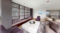 This elegant single-floor residence showcases a seamless blend of contemporary design and refined luxury. The expansive living area is anchored by floor-to-ceiling glass windows that wrap around the space, flooding the interior with natural light and offering sweeping panoramic views of the city skyline and waterfront. The living room features plush, neutral-toned sectional seating, a large upholstered ottoman, and a distinctive built-in bench with decorative backlit panels adorned with a floral motif in muted silver and lavender hues, adding an artistic focal point to the room.

Intricate parquet wood flooring in a geometric diamond pattern extends throughout the main living areas, contributing warmth and visual depth to the open-plan layout. A dedicated seating nook with custom built-in shelving with gold accents and dark stone paneling provides an intimate secondary lounge area. The kitchen is equally impressive, outfitted with richly veined stone countertops, custom wood cabinetry, premium stainless steel appliances, and a large central island — all framed by expansive city views through full-height glazing. A dining area with round table and upholstered seating sits adjacent, completing this sophisticated urban retreat.