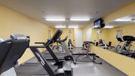 This fitness center is designed to provide a functional and well-equipped workout environment. The room features warm yellow walls and a neutral gray floor, creating a clean and energizing atmosphere. Bright fluorescent lighting installed across the ceiling ensures the space is well-lit throughout. A wall-mounted flat-screen television is positioned in the upper corner of the room, allowing users to stay entertained during their workouts.

The cardio section includes multiple treadmills lined along one side of the room, providing ample space for running or walking exercises. A multi-function cable machine and additional cardio equipment, such as a stationary bike, are also present, offering a variety of aerobic workout options. A rowing machine or flat bench is positioned centrally, adding to the versatility of the space.

Along one wall, a full-length mirror enhances the sense of space and allows users to monitor their form. Beneath the mirror, a dumbbell rack holds an organized selection of free weights in varying sizes. The room also features a door at the far end, likely providing access to additional facilities. Overall, this fitness center is efficiently arranged to support both cardio and strength training needs.