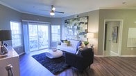 This single-floor residence presents a thoughtfully designed living area that balances modern aesthetics with everyday comfort. The space features rich hardwood flooring that runs throughout, providing a warm, cohesive foundation for the interior. A ceiling fan with an integrated light fixture is centrally positioned overhead, offering both functionality and a clean architectural detail.

The living room is anchored by a tufted white sofa and a dark accent chair, arranged around a contemporary oval coffee table atop a deep navy shag rug. A large-scale abstract artwork in muted greens, blues, and grays serves as a striking focal point on the main wall, adding visual depth and color to the space. A floor lamp nearby provides warm ambient lighting.

Natural light flows generously into the room through sliding glass doors that open directly onto an outdoor patio area, where outdoor seating is visible. The neutral gray walls complement the overall modern palette, while crown molding adds a refined architectural touch. A hallway visible along one side suggests additional rooms beyond the living area, contributing to the sense of a well-organized, functional floor plan.