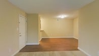 This single-floor space offers an open, versatile layout with a clean and neutral aesthetic throughout. The main living area is finished with warm hardwood flooring that adds a welcoming tone to the room, while a portion of the space transitions to light-colored carpeting, providing a subtle visual distinction between areas. The walls are painted in a soft, warm beige, creating a bright and inviting atmosphere throughout.

A flush-mount ceiling light provides overhead illumination, keeping the space well-lit. Visible HVAC venting in the ceiling indicates a central air system, contributing to year-round comfort. White baseboards run along the perimeter, offering a crisp contrast to the warm-toned walls and floors.

A pass-through or countertop cutout is partially visible along one wall, suggesting a connected kitchen or utility area nearby. A white door, likely leading to a closet or an adjacent room, is positioned near the entryway. Multiple electrical outlets are distributed along the walls, offering practical functionality.

Overall, the space presents a straightforward, move-in-ready environment with a neutral palette that allows for a wide range of interior design possibilities.