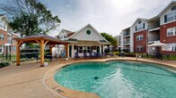 This outdoor amenity space centers around a generously sized freeform swimming pool with clearly marked depth zones, making it suitable for both leisurely swimming and relaxation. The pool's curvilinear design adds a resort-like quality to the area, complemented by clear blue water and a clean concrete surround that provides ample space for sunbathing and socializing.

Surrounding the pool, multiple lounge chairs are arranged along the deck, offering residents comfortable spots to relax and enjoy the open sky. A wood-framed gazebo with a pitched roof provides shaded seating and a gathering area, adding a warm, inviting architectural accent to the space.

Adjacent to the pool is a dedicated pool house, a white-sided structure with green-trimmed doors and a distinctive circular decorative detail, which likely houses restrooms and equipment. A wrought iron perimeter fence encloses the entire pool area, ensuring safety and privacy while maintaining clear sightlines to the surrounding landscaping and mature trees.

The broader setting includes multi-story residential buildings with brick and white siding facades, balconies, and green shutters, all of which frame the pool courtyard. Lush lawns and shade trees contribute to a tranquil, community-focused atmosphere, making this outdoor space an inviting retreat for residents year-round.