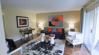 This single-floor living space showcases a thoughtfully designed interior that balances comfort and contemporary style. The main living area features warm, neutral-toned walls that serve as a backdrop for bold décor choices, including a striking two-panel abstract artwork in orange, gray, and black displayed prominently above a charcoal gray sofa accented with vibrant orange throw pillows. Dark hardwood flooring anchors the room, contrasting beautifully with a large abstract area rug in black and white tones. A pair of natural wood stump side tables and white pedestal tables serve as unique, organic coffee table accents at the center of the seating arrangement. A white upholstered accent chair with a slim metal side table adds a lighter visual element to the space. Ambient lighting is provided by matching table lamps on either side of the room, casting a warm glow throughout. A compact workspace or console desk area along one wall offers functional utility without compromising the open feel of the room. The overall ambiance is modern and curated, with a cohesive color palette of orange, gray, and neutral tones throughout.