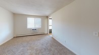 This single-floor unit at 335 Cowesett Ave in West Warwick, Rhode Island, offers a straightforward and functional layout designed for comfortable everyday living. The floor plan, as seen in the aerial view, organizes the living spaces efficiently, with carpeted rooms flowing logically from one to the next.

The living area is generously proportioned, featuring neutral-toned carpet, cream-colored walls, and a wall-mounted air conditioning unit positioned beneath a window that allows natural light to filter in. Baseboard heating is present throughout, providing a consistent heating solution. An adjacent room, visible through a doorway, appears to connect to an outdoor balcony area.

The bedroom includes a ceiling fan with a light fixture, a window with blinds offering natural light and outdoor views, and a sliding closet with mirrored doors and built-in rod storage. Baseboard heating is also present here.

The kitchen is equipped with white cabinetry, a gas range, a full-size refrigerator, and a double-basin sink beneath a window. Tile flooring and crown molding details give the kitchen a clean, organized appearance.

The bathroom features tile flooring, a full tub-shower combination, a vanity with storage, and a ventilation fan, completing this practical and well-maintained space.