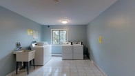 This single-floor laundry room is designed with functionality and practicality in mind. The space is finished with light blue walls that create a calm, clean atmosphere, complemented by white tile flooring that is easy to maintain. Multiple top-loading washers and front-loading dryers are arranged throughout the room, providing ample capacity for shared use. A utility sink stationed along one wall adds convenience for handwashing or pre-treating laundry. A centrally positioned window allows natural light to fill the room while offering a view of the surrounding greenery, reducing the reliance on the flush-mounted ceiling light. A baseboard heater runs along the wall beneath the window, ensuring comfort during cooler months. Informational notices are posted on the walls, keeping users informed of guidelines and procedures. The overall layout is open and well-organized, allowing for easy movement between machines. The space is clearly designed for shared, communal use, balancing efficiency with a tidy, welcoming environment.