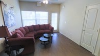 This single-story space features a well-lit living area that serves as its central gathering point. The room is finished with warm, hardwood-style flooring that runs throughout, adding a sense of continuity and warmth to the space. Neutral-toned walls provide a versatile backdrop, complementing a range of furnishing styles.

Natural light is a notable feature of the living area, provided by a large window along one wall and a glass-panel exterior door, which together create a bright and open atmosphere. A ceiling fan with an integrated light fixture is centrally mounted overhead, offering both air circulation and supplemental lighting.

The layout comfortably accommodates a large sectional sofa and a circular glass-top coffee table, demonstrating the room's capacity for practical furniture arrangements. Electrical outlets are conveniently positioned along the walls, and clean baseboards frame the perimeter of the room. The exterior door provides direct access to the outdoors, enhancing the functionality of the living space. Overall, the area presents a clean, functional, and welcoming environment.