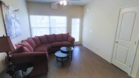 This single-story space features a well-lit living area that serves as its central gathering point. The room is finished with warm, hardwood-style flooring that runs throughout, adding a sense of continuity and warmth to the space. Neutral-toned walls provide a versatile backdrop, complementing a range of furnishing styles.

Natural light is a notable feature of the living area, provided by a large window along one wall and a glass-panel exterior door, which together create a bright and open atmosphere. A ceiling fan with an integrated light fixture is centrally mounted overhead, offering both air circulation and supplemental lighting.

The layout comfortably accommodates a large sectional sofa and a circular glass-top coffee table, demonstrating the room's capacity for practical furniture arrangements. Electrical outlets are conveniently positioned along the walls, and clean baseboards frame the perimeter of the room. The exterior door provides direct access to the outdoors, enhancing the functionality of the living space. Overall, the area presents a clean, functional, and welcoming environment.