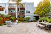 Located at 222 Columbia St in Santa Cruz, California, this beautifully designed outdoor courtyard serves as a central gathering space surrounded by multi-story residential buildings with warm wood-railed balconies and a contemporary gray exterior. The ground level is finished with broad, natural stone pavers in earthy tones that lend a polished yet relaxed coastal character to the space.

Mature palm trees rise prominently throughout the courtyard, anchoring the landscape and contributing a distinctly Californian atmosphere. Thoughtfully designed garden beds line the perimeter, filled with tropical foliage, ornamental grasses, and colorful plantings that provide both privacy and visual interest. Stone retaining walls border several of the planted areas, adding a structured architectural element to the lush greenery.

Multiple seating arrangements are distributed across the courtyard to accommodate a variety of uses. A modern lounge area features a round white coffee table, a sofa with accent pillows, and wire-frame accent chairs. A casual dining area with a long bench table offers a spot for outdoor meals, while additional seating clusters invite conversation throughout the space. Overhead string lights are strung across the courtyard, creating a warm, inviting ambiance after dark. A pergola with an outdoor grill station is visible toward the rear, and pool access is available nearby, rounding out this well-appointed community outdoor environment.