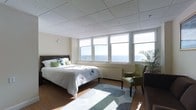 This inviting space offers a thoughtfully designed single-floor layout that seamlessly blends comfort with coastal beauty. The room is anchored by a generously sized bed dressed in crisp white linens and accented with green decorative pillows, paired with a classic dark wood headboard that adds a touch of warmth. A stylish green accent chair with nailhead trim provides an elegant spot for reading or unwinding, complemented by a lush potted palm that brings a touch of nature indoors. The warm hardwood floors are softened by a contemporary area rug in muted gray and white tones, adding texture and visual interest to the space. A suspended grid ceiling with recessed panels lends the room a clean, modern feel while allowing ample overhead space. The most striking feature is the wide bank of windows along the exterior wall, framing breathtaking panoramic ocean views that flood the room with natural light throughout the day. Roller shades offer privacy and light control as needed. The overall ambiance is calm and airy, with a coastal-inspired palette of whites, greens, and natural wood tones that evoke a sense of tranquility and escape.