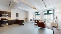 This single-floor urban loft blends industrial character with contemporary design across a thoughtfully arranged open floor plan. The expansive main living area is defined by soaring ceilings with exposed cylindrical ductwork and track lighting, lending an authentic loft aesthetic. Polished concrete-style floors run throughout, providing a clean, cohesive foundation for the entire space.

Large, boldly green-framed windows dominate the living and dining area, flooding the interior with natural light while offering striking views of the surrounding cityscape. The living zone features a comfortable leather seating arrangement anchored by a patterned area rug, alongside a media console and coffee table. The dining area accommodates a round table with modern chairs, positioned to take full advantage of the window views.

The kitchen is equipped with dark espresso cabinetry, stainless steel appliances including a refrigerator and dishwasher, a white subway tile backsplash, and a light stone countertop. A peninsula with bar seating provides an additional casual dining option.

The bedroom offers a quieter, more intimate setting with vaulted beam detailing at the ceiling, double closets, and a serene, neutral palette. The bathroom includes a full tub-shower combination with subway tile surround, a granite-topped vanity, and clean, functional finishes throughout.