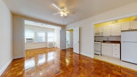 This single-floor residence showcases rich, honey-toned parquet hardwood flooring throughout, lending warmth and classic character to every room. The open living area is the centerpiece of the space, featuring a ceiling fan with light fixture and a wide arched entryway that connects seamlessly to an additional room, creating a generous, flowing layout. Natural light filters in through multiple windows, each equipped with a window-mounted air conditioning unit, keeping the space comfortable year-round. A radiator along the wall provides supplemental heating. The kitchen, distinguished by bright yellow accent walls, is efficiently designed with white cabinetry, tile backsplash, a gas range, microwave, and a full-size refrigerator — offering all essential appliances in a compact footprint. The bedroom is a calm retreat with parquet floors continuing underfoot, large windows with blinds, and direct access to a closet. The bathroom is finished in a timeless black-and-white tile scheme, complete with a full bathtub, pedestal sink, toilet, and a small window that allows natural light. Throughout the unit, neutral cream walls provide a clean, versatile backdrop that complements the warm flooring and traditional architectural details.
