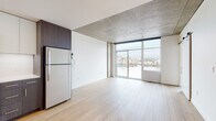 This contemporary single-floor unit showcases a thoughtfully designed open-concept layout that seamlessly connects the living, dining, and kitchen areas. The exposed concrete ceiling serves as a defining architectural feature, lending an industrial-modern character that is complemented by smooth white walls and light-toned hardwood flooring throughout.

The living area is anchored by expansive floor-to-ceiling windows that flood the space with natural light and frame impressive views of a nearby waterway and bridge, creating a striking visual focal point. A barn-style sliding door adds a stylish, space-efficient detail to the interior.

The kitchen is well-appointed with a full suite of stainless steel appliances, including a gas range, built-in microwave, dishwasher, and a full-size refrigerator. Two-tone cabinetry — dark wood lower cabinets paired with white upper cabinets — provides ample storage while contributing to the modern aesthetic. A clean white countertop and under-cabinet lighting complete the functional and polished look.

The bathroom features a full soaking tub and shower combination enclosed in glossy subway tile, with clean white fixtures and a neutral tile floor that maintain the unit's crisp, contemporary feel. Overall, the space offers a sleek, urban living environment with high-quality finishes and an abundance of natural light.