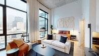 Located in Chicago's vibrant South Loop neighborhood, this thoughtfully designed studio at 633 South LaSalle Street offers a compelling blend of industrial character and contemporary finishes. The space is defined by its dramatic exposed concrete ceiling and expansive floor-to-ceiling windows framed in sleek black steel, flooding the interior with natural light and offering striking views of the surrounding cityscape.

The open-plan main area seamlessly integrates the living, dining, and kitchen zones. Light wood-tone flooring runs throughout, providing warmth that balances the raw ceiling above. The kitchen is outfitted with dark, wood-grain cabinetry extending to ceiling height, stainless steel appliances including a refrigerator, dishwasher, and range, and a clean white countertop. A sliding barn-style door adds a modern, space-conscious touch to the layout.

The living area accommodates a comfortable seating arrangement alongside a dedicated sleeping area, all oriented to take full advantage of the city views. A dedicated closet provides organized storage, while the bathroom features a clean, minimal aesthetic with light wood-look flooring, open shelving, and crisp white walls. In-unit laundry adds everyday convenience, completing this efficient and stylish urban residence.