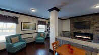 Located at 8444 Easton Commons Dr in Louisville, Kentucky, this single-floor communal space is thoughtfully designed to offer both comfort and functionality. The interior is unified by rich, dark hardwood flooring and striking dark crown molding that frames the cream-toned walls, creating a polished and cohesive aesthetic throughout.

At the heart of the space stands a prominent stone-clad fireplace with a sleek built-in linear firebox, topped by a wall-mounted flat-screen television. The stacked stone surround extends from floor to ceiling, serving as a dramatic focal point that anchors a cozy seating area furnished with upholstered accent chairs. A decorative structural column with dark trim detailing adds architectural character and subtly defines the transition between zones.

The dining area features multiple wooden tables with ladder-back chairs, providing ample seating for group gatherings. Natural light filters in through windows dressed with dark grommet-top curtains, lending a warm and inviting atmosphere. A built-in shelving unit adds functional display space, while framed artwork, including a vibrant floral print, contributes a lively touch of color.

A hallway connects to adjacent areas, accented by pendant lighting and a large decorative wall clock. The granite-topped counter along the stone base adds an elegant finishing detail. Overall, the space exudes a warm, community-oriented ambiance well-suited for social interaction and relaxation.