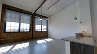 This single-floor loft showcases a compelling blend of industrial heritage and modern design. The open-plan layout is anchored by striking exposed brick walls that add warmth and texture, complemented by soaring ceilings with raw, unfinished detailing that preserve the character of the original structure. Overhead, black pipe conduit runs visibly along the ceiling, reinforcing the industrial aesthetic, while pendant lighting fixtures hang at intervals to provide functional illumination with a stylish edge.

The polished concrete floors reflect the abundant natural light that pours in through the large, multi-pane factory-style windows fitted with black metal frames. These generously sized windows stretch nearly floor to ceiling and offer open views of the surrounding exterior, making the space feel bright and connected to the outdoors. The combination of light-toned walls and reflective flooring enhances the sense of openness throughout.

Along one side of the space, a kitchen area is partially visible, featuring a granite countertop peninsula that provides both workspace and casual seating. The cabinetry below features rich wood tones that contrast with the otherwise industrial palette, adding a refined touch. Overall, this space delivers a distinctive atmosphere that balances raw, historic character with clean, contemporary finishes.
