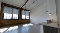 This single-floor loft showcases a compelling blend of industrial heritage and modern design. The open-plan layout is anchored by striking exposed brick walls that add warmth and texture, complemented by soaring ceilings with raw, unfinished detailing that preserve the character of the original structure. Overhead, black pipe conduit runs visibly along the ceiling, reinforcing the industrial aesthetic, while pendant lighting fixtures hang at intervals to provide functional illumination with a stylish edge.

The polished concrete floors reflect the abundant natural light that pours in through the large, multi-pane factory-style windows fitted with black metal frames. These generously sized windows stretch nearly floor to ceiling and offer open views of the surrounding exterior, making the space feel bright and connected to the outdoors. The combination of light-toned walls and reflective flooring enhances the sense of openness throughout.

Along one side of the space, a kitchen area is partially visible, featuring a granite countertop peninsula that provides both workspace and casual seating. The cabinetry below features rich wood tones that contrast with the otherwise industrial palette, adding a refined touch. Overall, this space delivers a distinctive atmosphere that balances raw, historic character with clean, contemporary finishes.