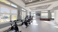 Located at 1450 Neo Square Blvd in Kissimmee, Florida, this modern fitness studio offers a well-appointed, single-floor layout designed to support a variety of workout routines. The space is divided into two connected areas, each thoughtfully designed with a cohesive gray-and-white color palette and light-toned luxury vinyl flooring throughout.

The primary cycling room houses a row of stationary spin bikes equipped with individual screens, positioned alongside a bank of large windows that flood the space with natural sunlight and offer views of the surrounding outdoors. A decorative sculptural figure adds an artistic touch to the room's clean, contemporary aesthetic. Coffered ceilings with recessed lighting and ceiling fans enhance both the visual appeal and comfort of this area.

An adjoining open exercise studio provides ample floor space for stretching, group fitness, or strength training. Full wall-to-wall mirrors amplify the sense of space and allow users to monitor their form during workouts. A large mounted flat-screen television serves as a focal point, while wooden benches along the walls offer convenient seating. The room also features a well-stocked rack of exercise balls, resistance tools, and fitness accessories. High windows along multiple walls ensure continuous natural light, creating an energizing and inviting atmosphere throughout the entire facility.
