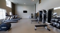 This fitness center offers a well-appointed and thoughtfully organized exercise space designed to accommodate a variety of workout routines. The interior features a clean, minimalist aesthetic with neutral-toned walls and a smooth, durable flooring surface that provides a practical foundation for physical activity. Recessed ceiling lighting ensures the space is evenly and brightly lit throughout.

The cardio zone is anchored by a row of treadmills positioned along one side of the room, offering ample space for users to move freely. Toward the center and far end of the space, a selection of strength and resistance training machines are arranged to allow efficient use of the floor area. A flat-panel television is mounted on the wall, providing entertainment for those engaged in longer cardio sessions. Flat workout benches are also available, complementing the weight and resistance machines for a versatile strength training experience.

The open layout of the fitness center encourages ease of movement between stations without feeling crowded. High ceilings contribute to an airy, spacious ambiance, making the environment feel comfortable even during peak use. Overall, this fitness center is a functional and modern facility well-suited for a comprehensive daily workout regimen.