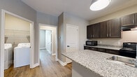 Located at 4950 Weaver Rd in Lake Charles, Louisiana, this single-story, 5-bedroom, 2-bathroom home offers a well-designed layout with modern finishes throughout. The open-concept kitchen and living area serves as the heart of the home, showcasing a large center island with granite countertops, dark espresso cabinetry, pendant lighting, and black appliances including a range and refrigerator. Wood-look luxury vinyl flooring flows consistently throughout every room, complementing the warm, neutral gray wall tones and bright white trim and baseboards. Each of the five bedrooms is well-proportioned, featuring natural light from windows dressed with white blinds and ceiling fans in select rooms. Closets with wire shelving provide practical storage. The two bathrooms are finished with granite-topped dark vanities, large mirrors, and tub-shower combinations. A dedicated in-unit laundry room with full-size washer and dryer hookups adds everyday convenience. A central hallway connects the rooms efficiently, and the overall layout—visible in the dollhouse view—reflects a thoughtful, functional design. Clean lines, consistent finishes, and a neutral palette give this home a move-in-ready appeal suited for comfortable everyday living.