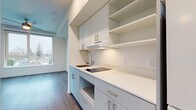 This single-floor unit showcases a clean, modern design characterized by neutral white walls, warm wood-tone luxury vinyl plank flooring, and recessed lighting throughout. The layout flows efficiently from a central hallway connecting the kitchen and living areas to the individual rooms.

The kitchen is outfitted with sleek light-gray cabinetry featuring brushed nickel hardware, a white quartz countertop, an undermount sink with a contemporary faucet, and a built-in cooktop. Open upper shelving complements the upper cabinets, adding both storage and visual interest. A range hood is integrated seamlessly into the cabinetry design.

The living and sleeping areas are bright and open, each benefiting from large windows that frame outdoor views of the surrounding neighborhood and green spaces. A ceiling fan is present in the main living area. One room features a sliding barn-style door, adding a modern design detail. A compact room with a window provides flexible use as a den or storage space.

The bathroom is finished in a minimalist style with wood-look flooring, chrome fixtures, and clean white walls. Overall, the unit presents a contemporary and functional aesthetic suited for everyday living.