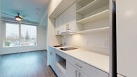 This single-floor unit showcases a clean, modern design characterized by neutral white walls, warm wood-tone luxury vinyl plank flooring, and recessed lighting throughout. The layout flows efficiently from a central hallway connecting the kitchen and living areas to the individual rooms.

The kitchen is outfitted with sleek light-gray cabinetry featuring brushed nickel hardware, a white quartz countertop, an undermount sink with a contemporary faucet, and a built-in cooktop. Open upper shelving complements the upper cabinets, adding both storage and visual interest. A range hood is integrated seamlessly into the cabinetry design.

The living and sleeping areas are bright and open, each benefiting from large windows that frame outdoor views of the surrounding neighborhood and green spaces. A ceiling fan is present in the main living area. One room features a sliding barn-style door, adding a modern design detail. A compact room with a window provides flexible use as a den or storage space.

The bathroom is finished in a minimalist style with wood-look flooring, chrome fixtures, and clean white walls. Overall, the unit presents a contemporary and functional aesthetic suited for everyday living.